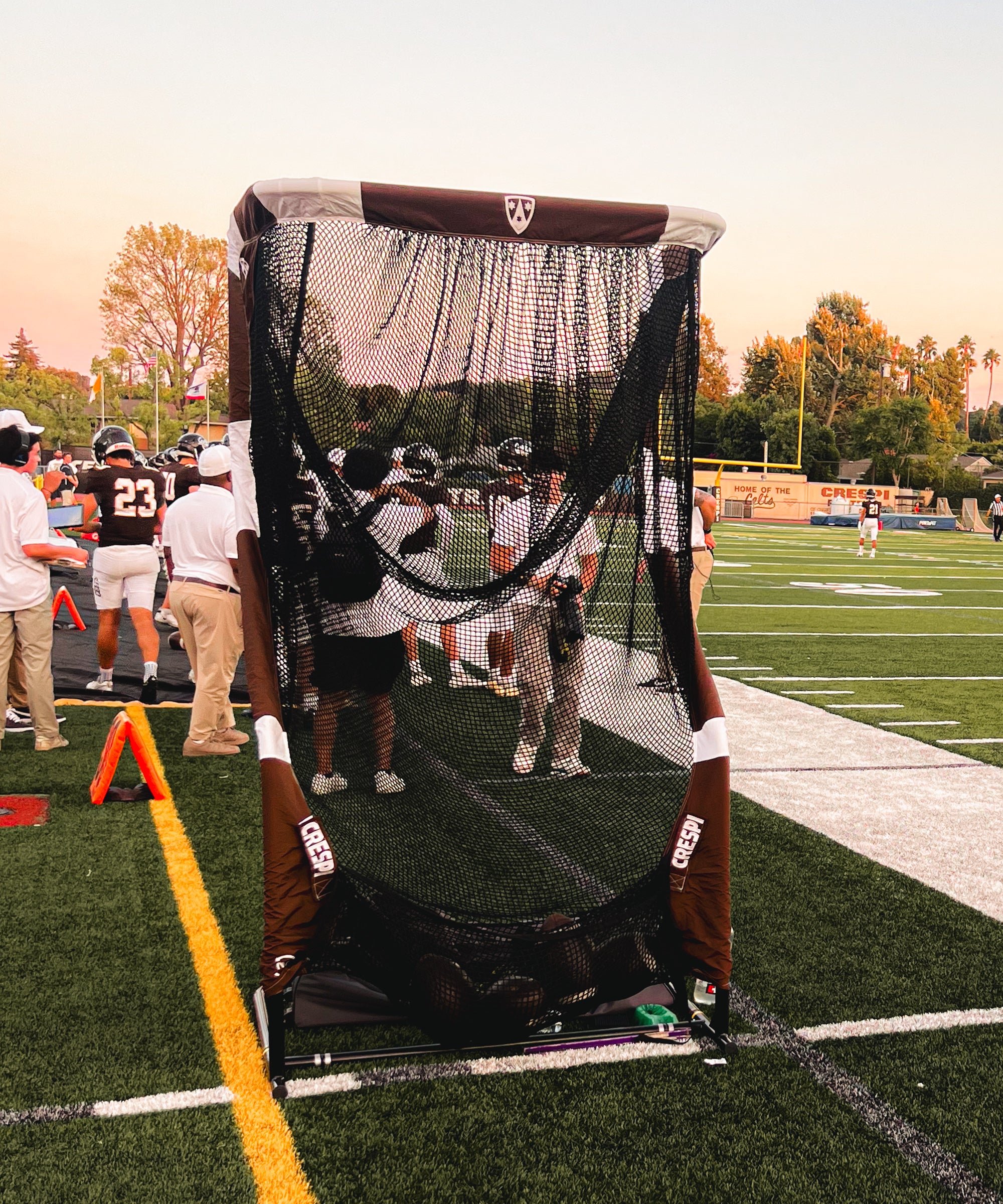 Football practice net on a field with players and coaches in the background.
