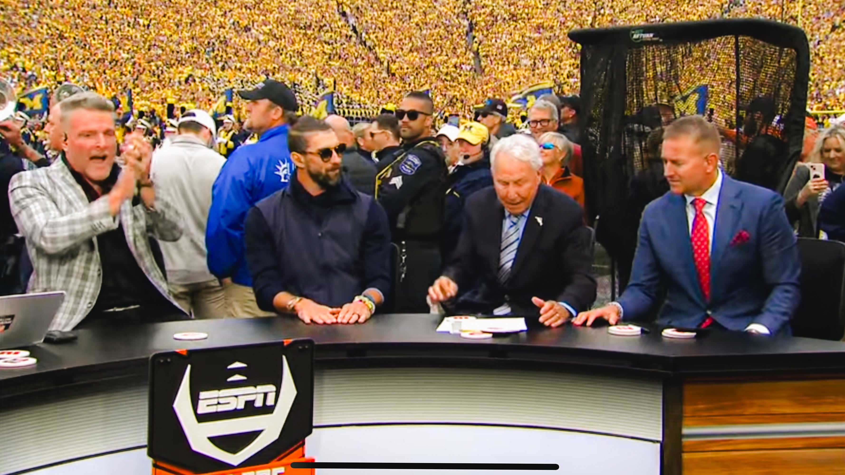 Four men sit at an ESPN desk outdoors, with a large crowd in yellow behind them. One man is clapping, another is wearing sunglasses.