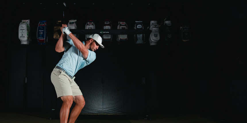 A golfer in a blue shirt and beige shorts prepares to swing indoors, with a row of golf bags displayed in the background.