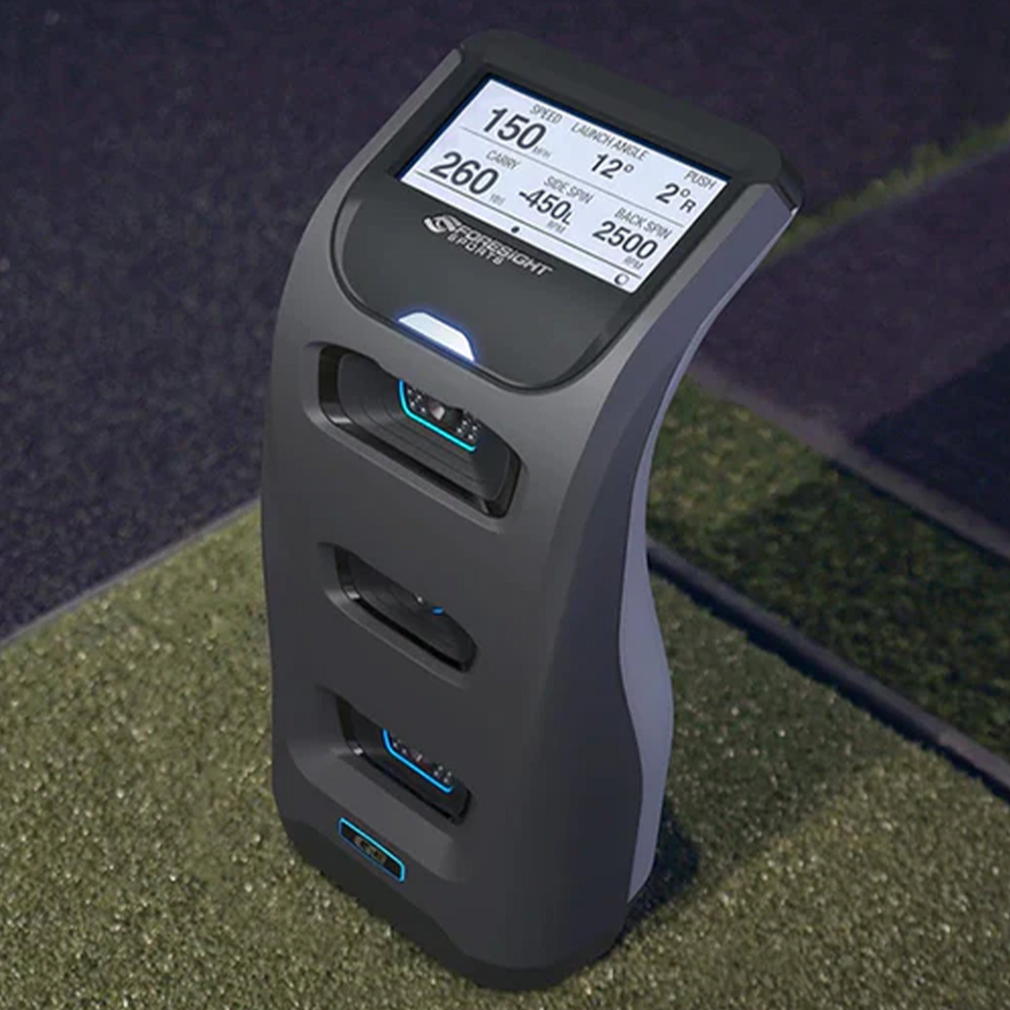 A golf launch monitor with a digital display showing swing data, positioned on a patch of grass.