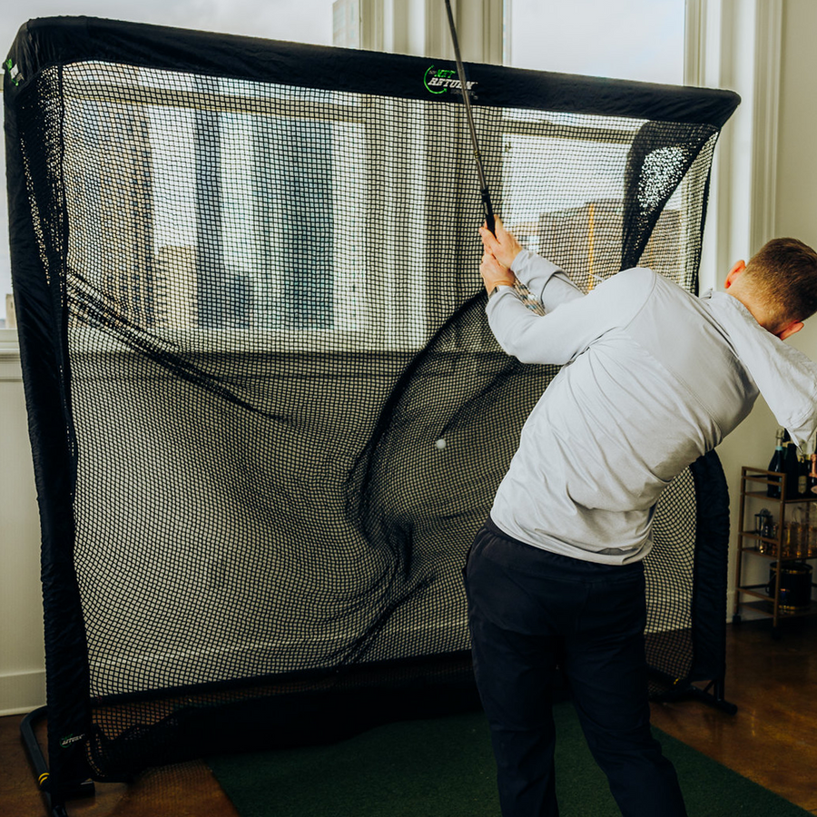 A person in a light hoodie practices a golf swing indoors using a net.