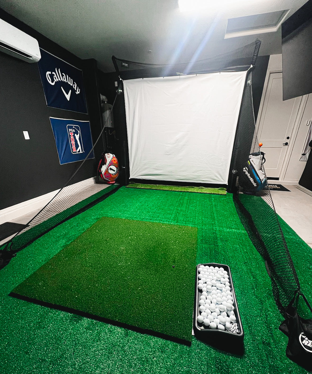 Indoor golf simulator setup with a hitting mat, practice net, and a container of golf balls. Wall banners in the background.