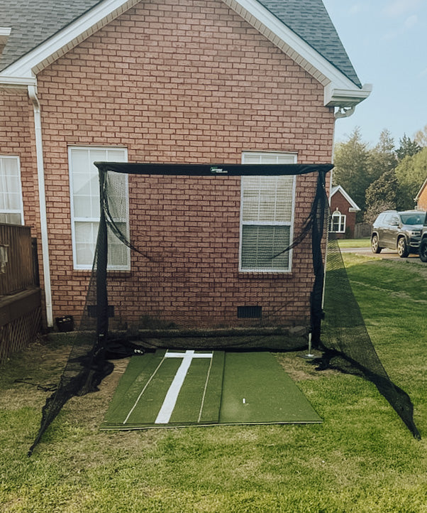 A golf practice net set up on the lawn next to a brick house, with a green mat and a white target line.