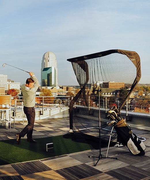 Person practicing golf swings on a rooftop with a net, overlooking a cityscape featuring modern buildings.