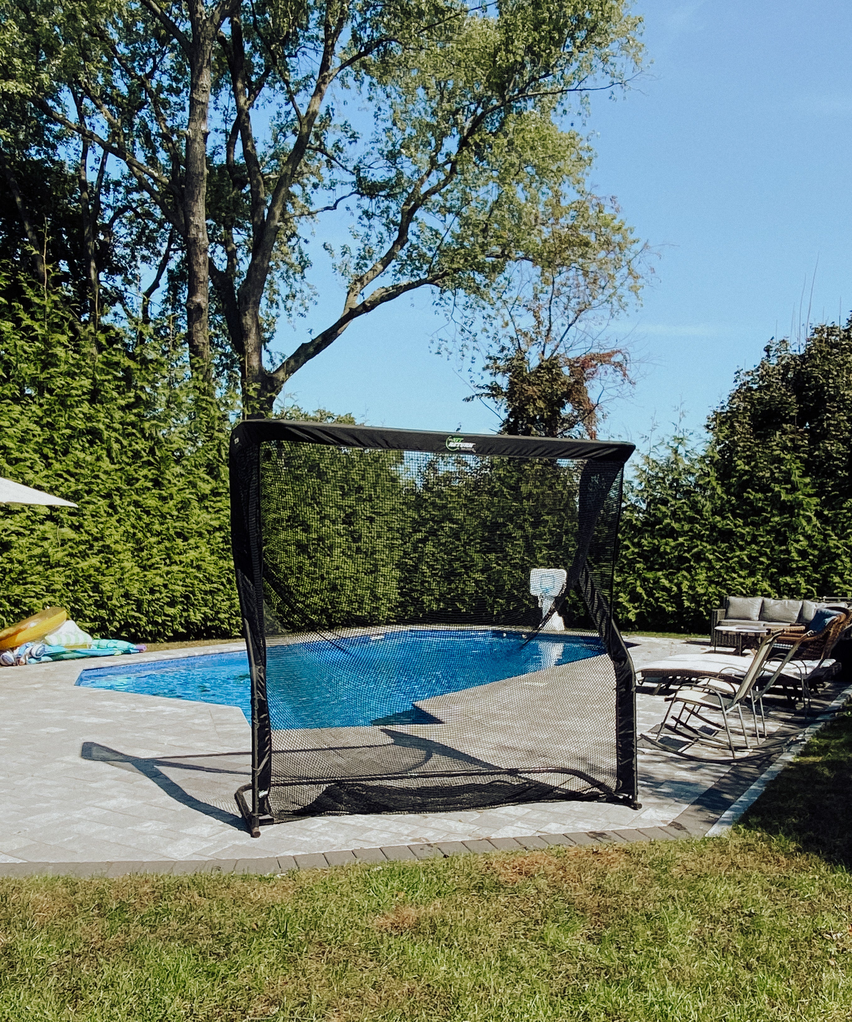A portable soccer goal stands near a swimming pool surrounded by patio chairs and trees under a clear sky.