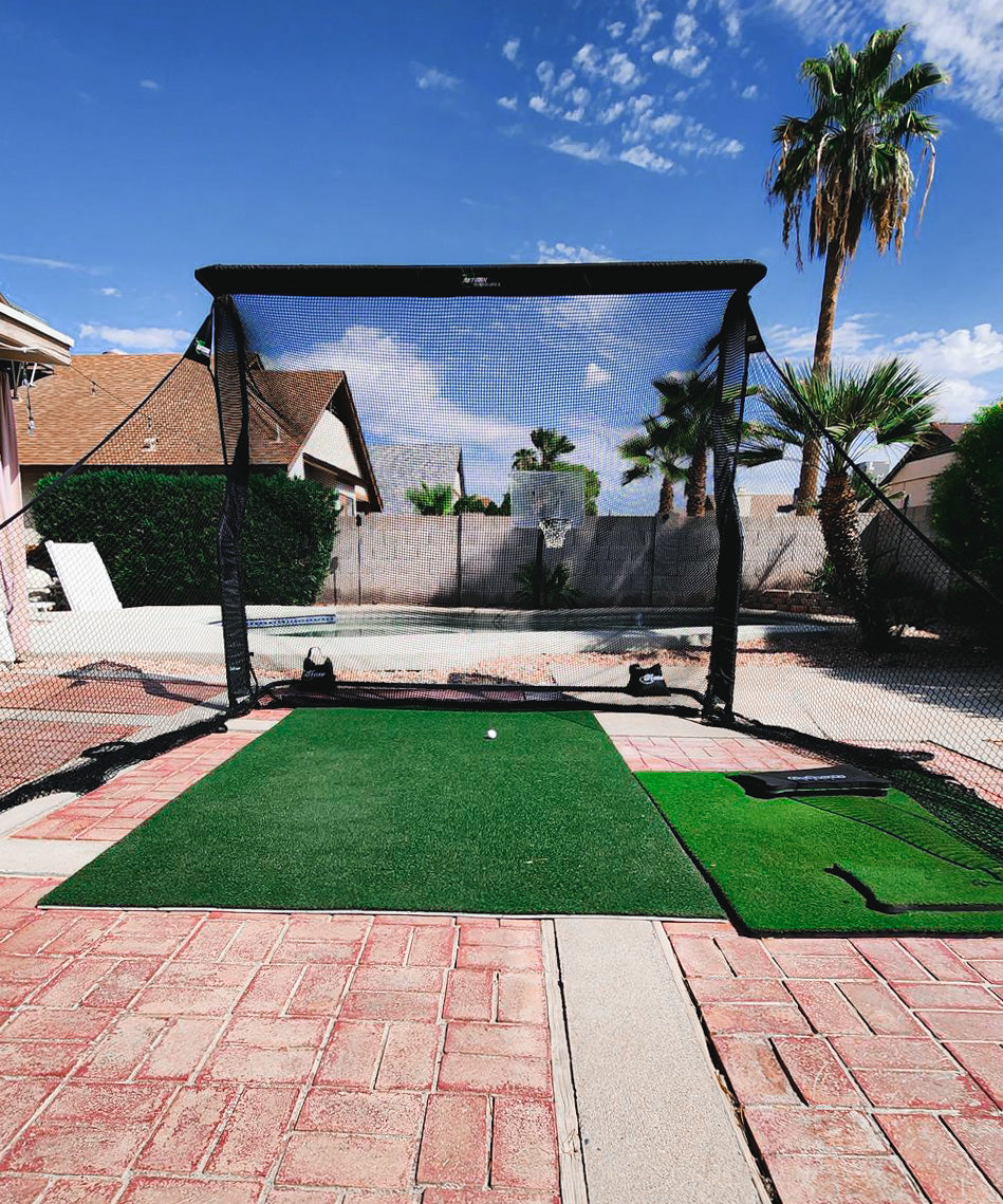 A backyard golf practice net set up on a brick patio with artificial turf, surrounded by palm trees under a clear blue sky.