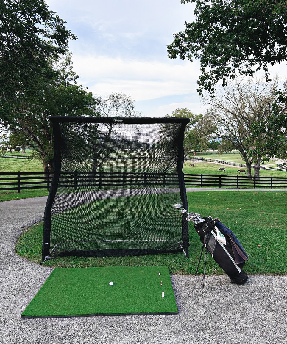 Golf practice setup with a net, mat, and club bag on a paved path. Green field and fence in the background.