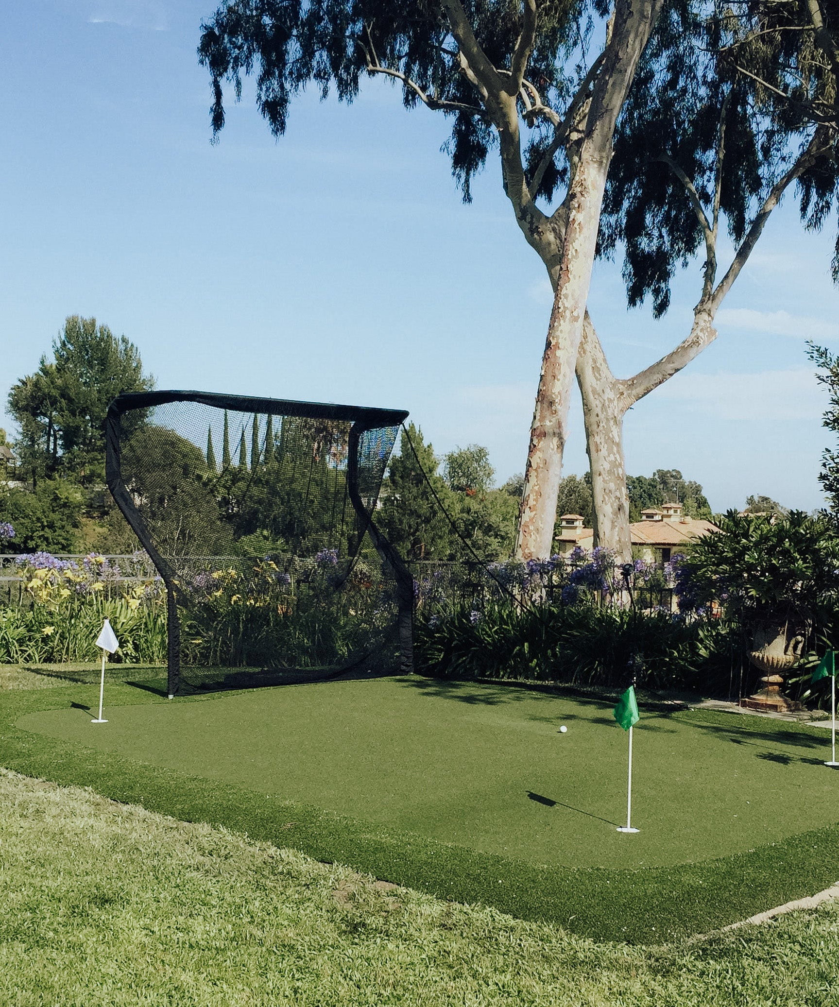 A small outdoor putting green with two flags and a golf net, surrounded by trees and greenery under a clear blue sky.
