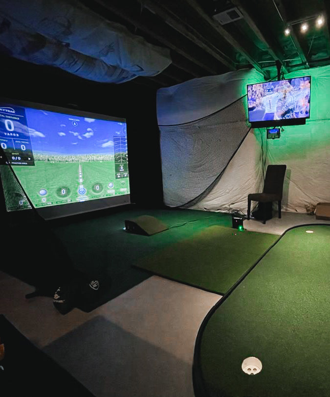 Indoor golf simulator setup with screen displaying a golf course, putting green, chair, and netting. A TV above shows a sporting event. Room has exposed ceiling and dim lighting.
