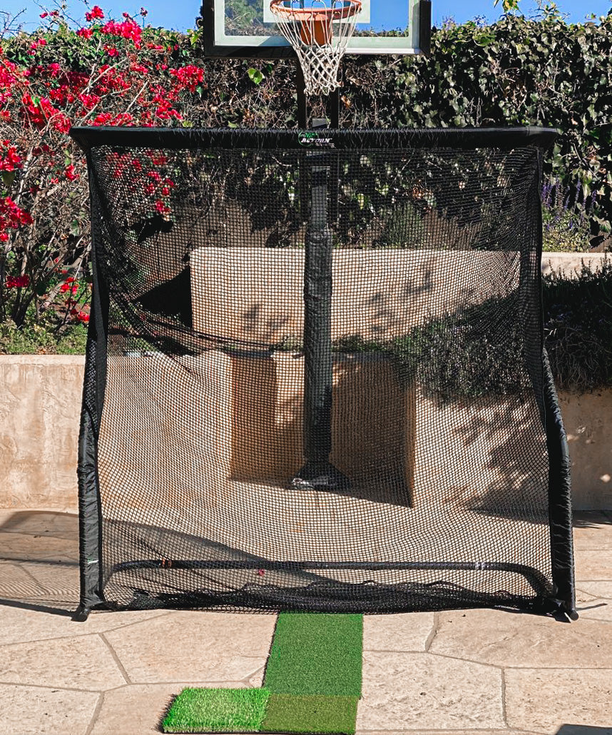 A black net rebounder positioned on a stone patio, with a basketball hoop and red flowers in the background.