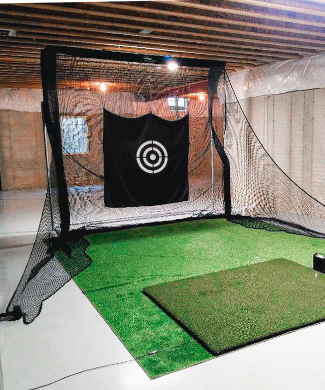 Indoor golf practice net with target, set up on a green mat in a basement with unfinished walls and exposed wood beams.