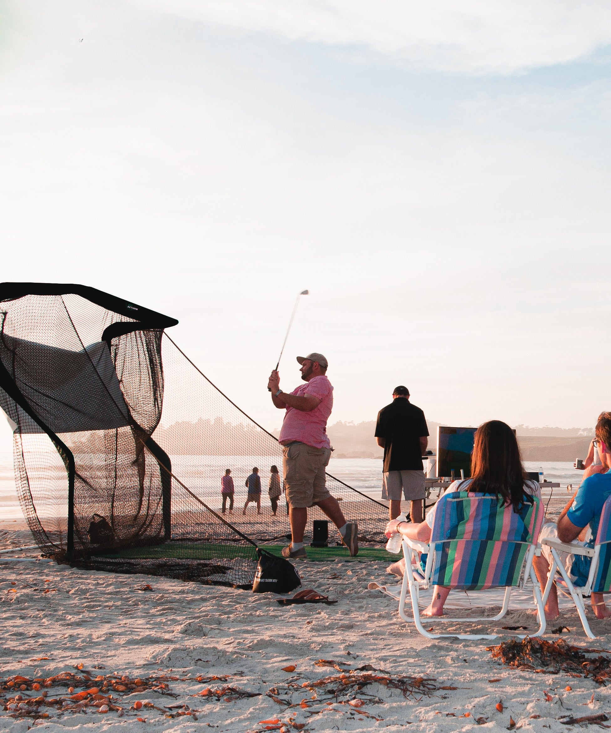 A man swings a golf club on a beach using a practice net, while seated people observe.