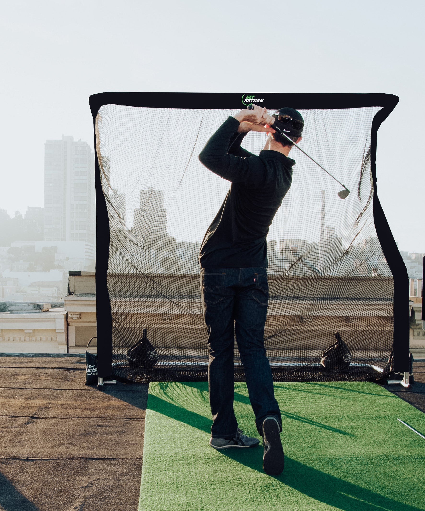 A person practices golf swings on a rooftop with a hitting net and mat. Buildings are visible in the background.