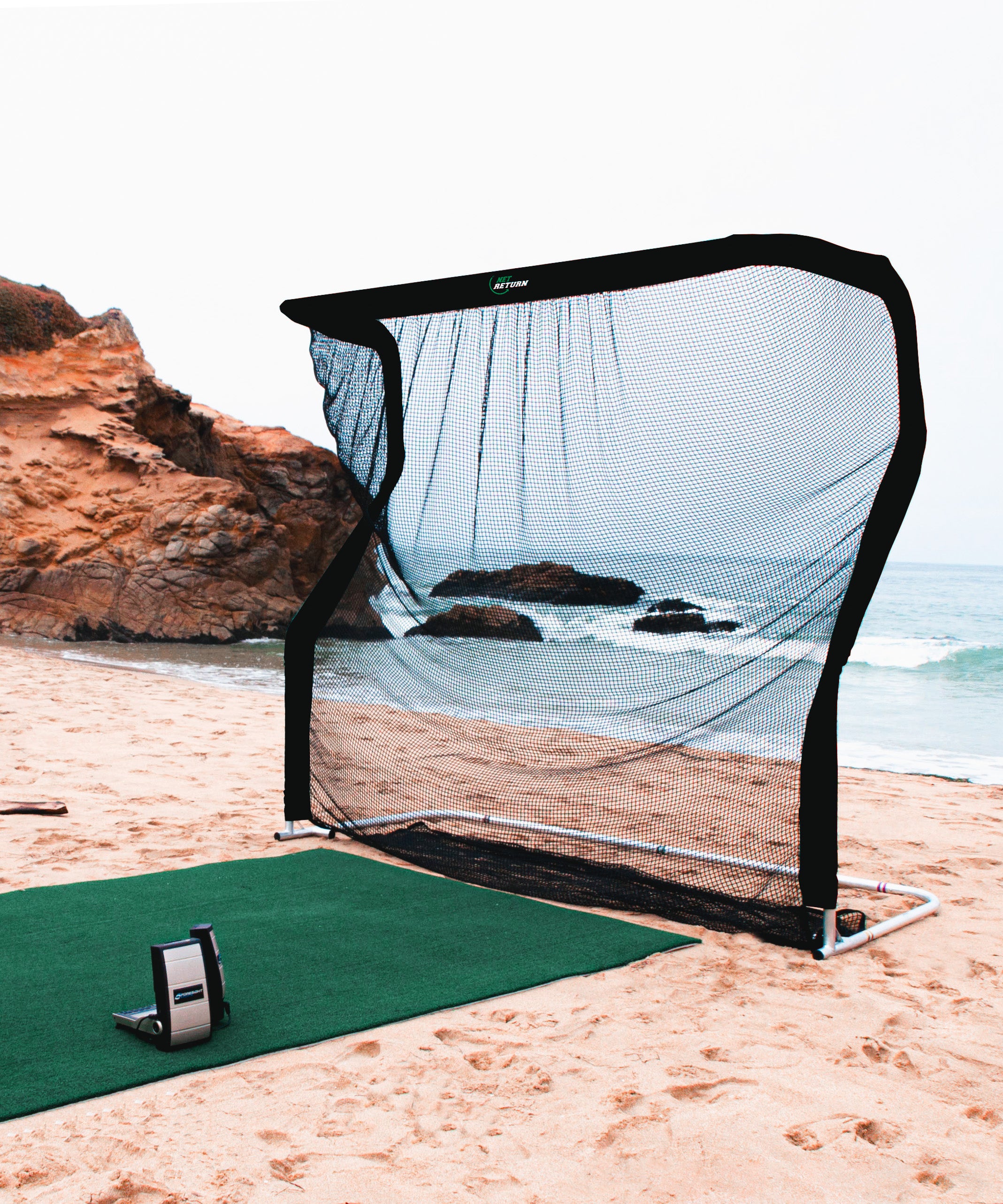 A golf practice net set up on a beach, with a turf mat and rocky coastline in the background.