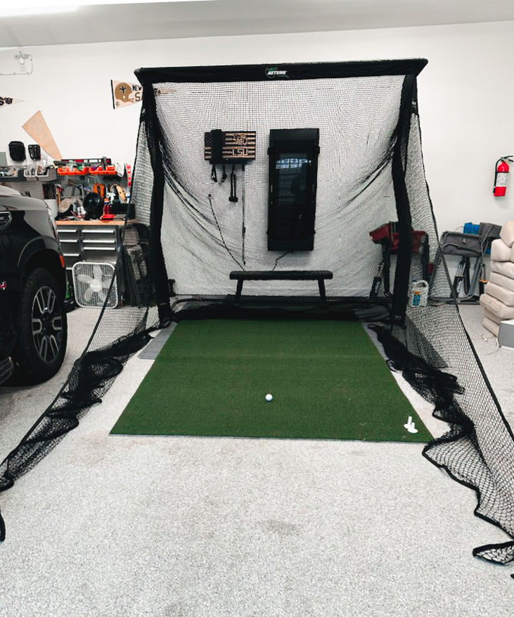Indoor golf practice setup with a net, mat, and golf ball in a garage. Tools and a fire extinguisher are visible on the walls.