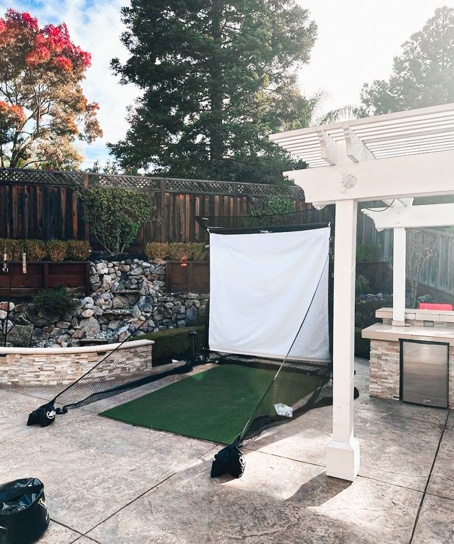 A backyard golf practice setup with a hitting net and mat on a concrete patio. There's a wooden pergola and landscaping with trees and rocks in the background.
