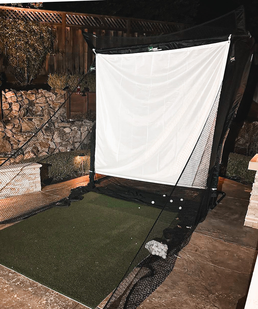 A golf practice net set up in a backyard, with a mat and several golf balls nearby. Stone wall and wooden fencing in the background.
