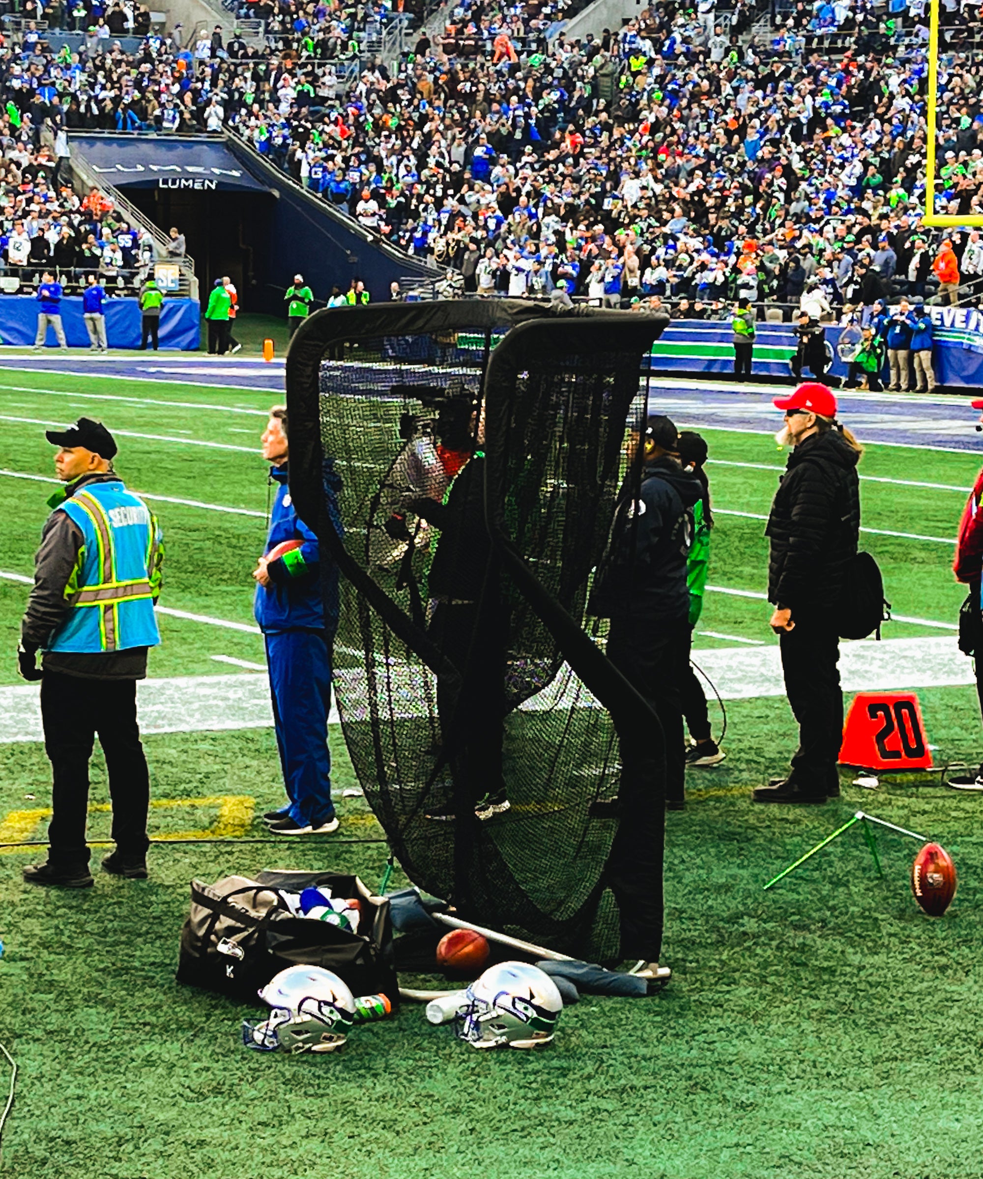 A football player practices on the sidelines with a net during a game. Helmets, a football, and staff are visible around the field.