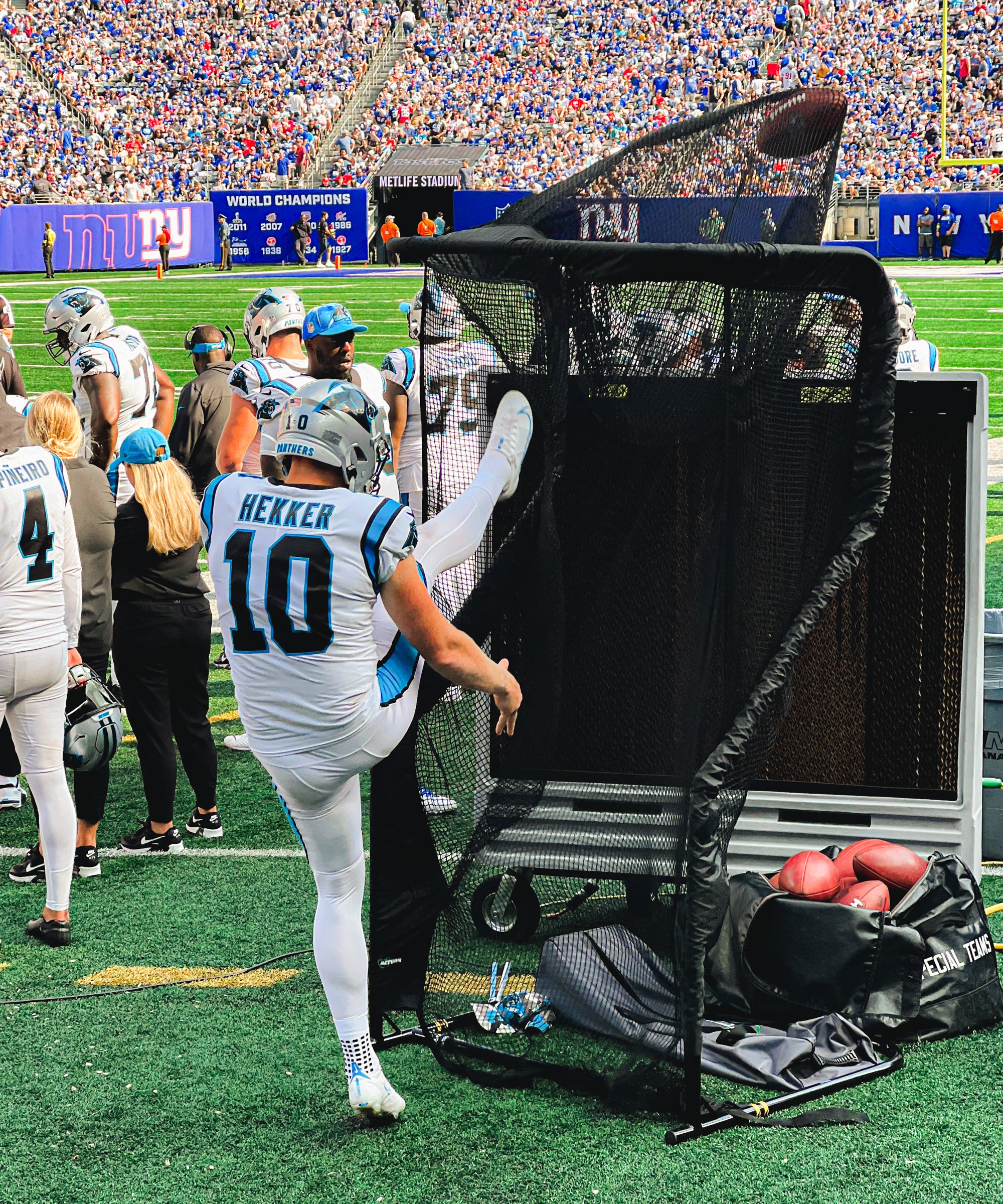 Football player in a white uniform practices punting into a net on the sideline during a game. Crowd and teammates are visible in the background.