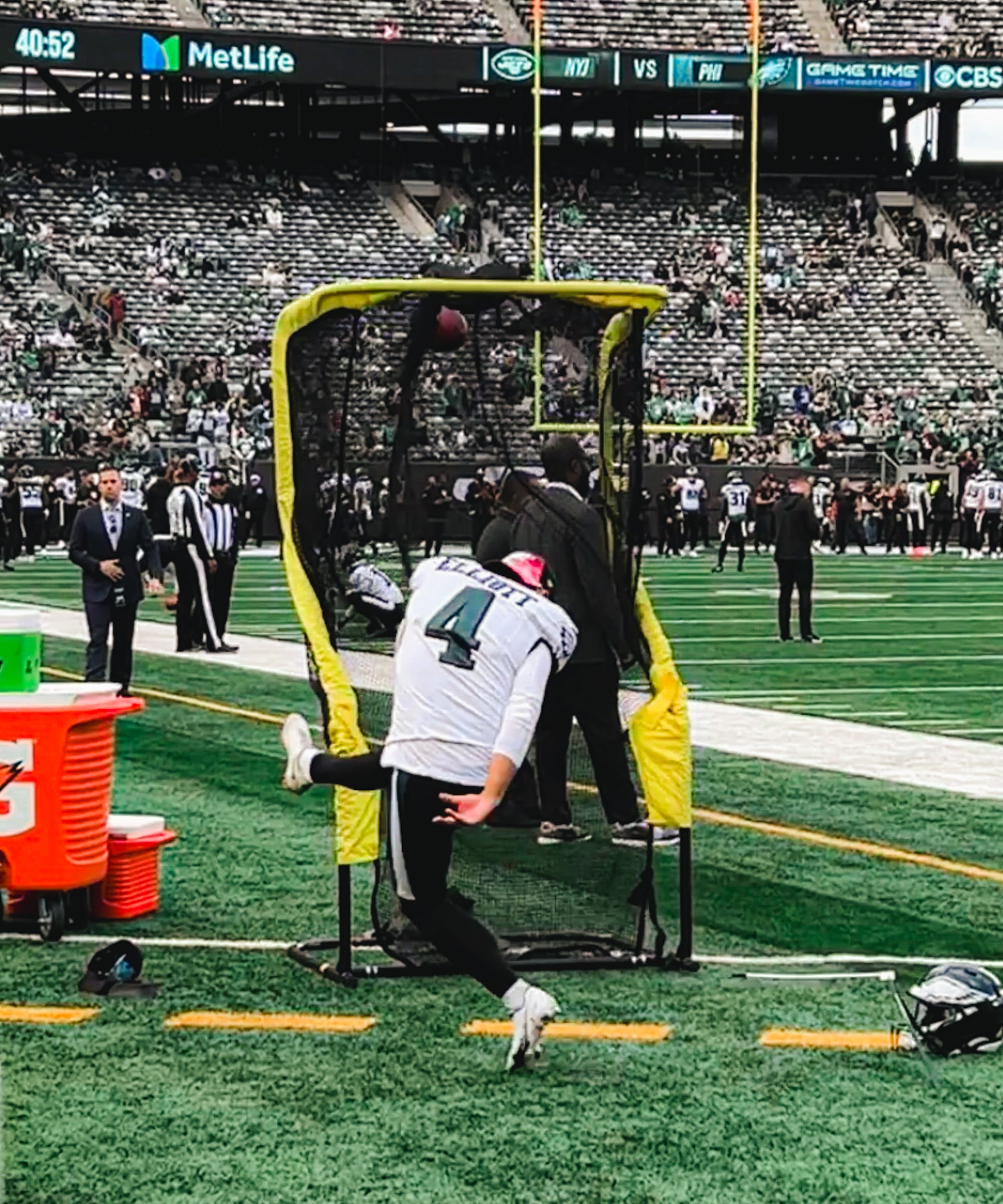 Football player practicing kicking into a net on a field with people and a stadium in the background.