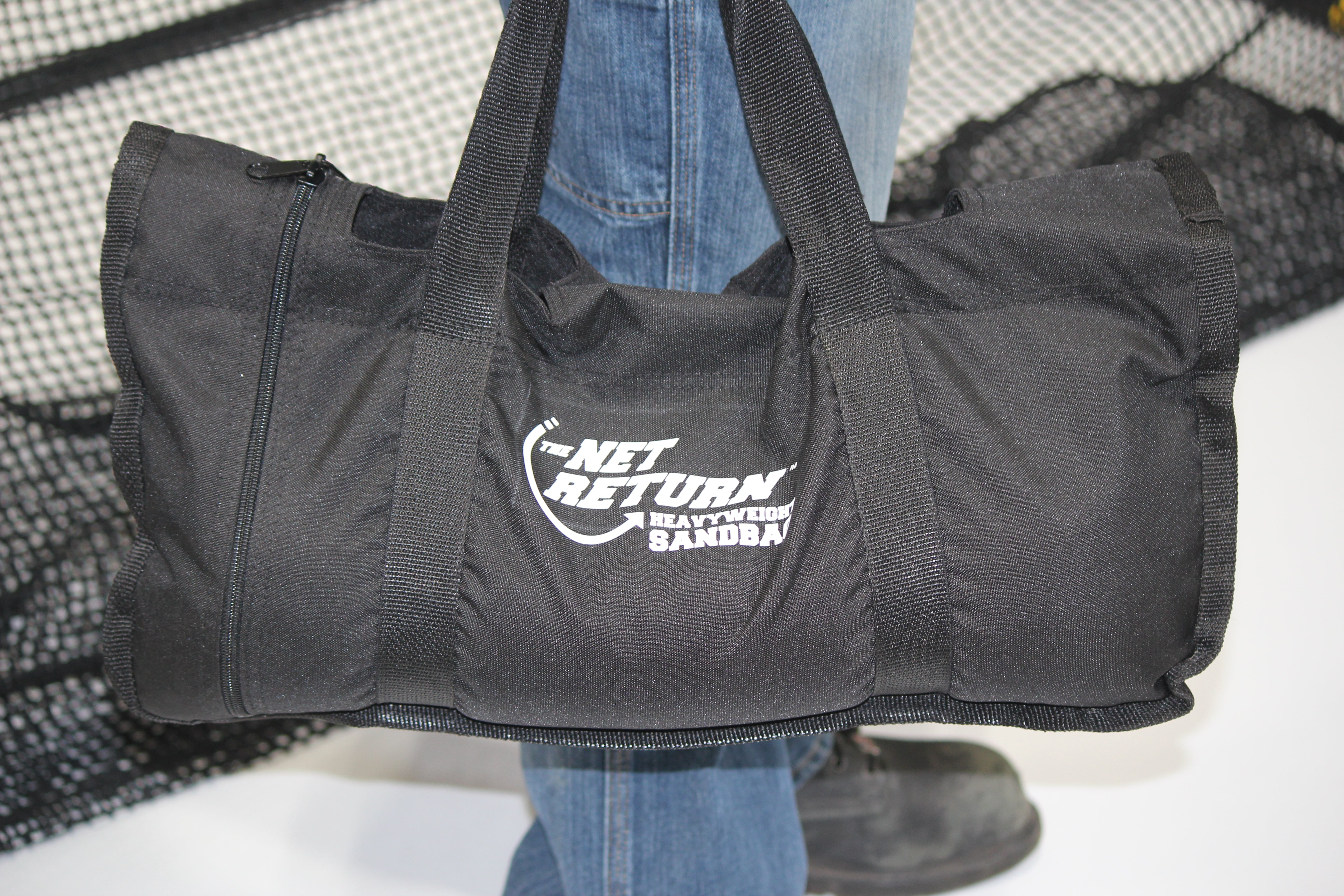 A person holds a black duffel bag labeled "The Net Return Heavyweight Sandbag" with handles and a zipper.