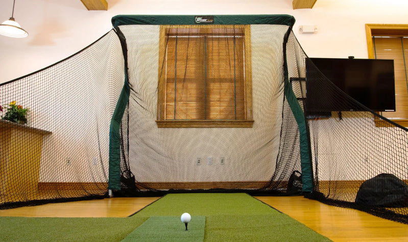 Indoor golf practice setup with a hitting net, green mat, and golf ball placed on a tee in a room with wooden floors and window blinds.